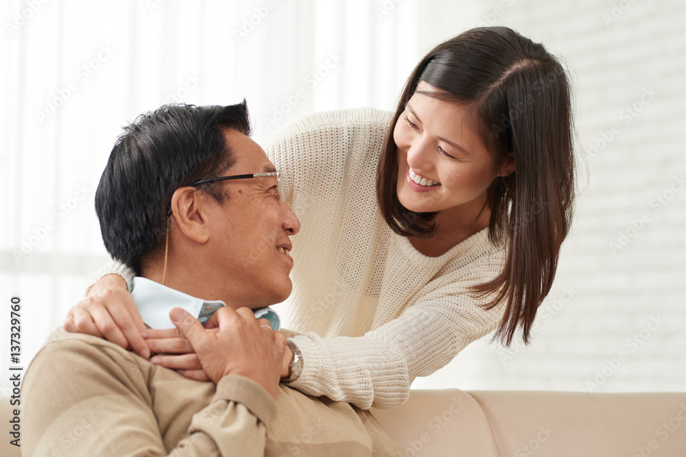 Lovely family moments: Asian young woman standing behind her senior dad and hugging him tenderly, he turned to her and smiling Lovely family moments: Asian young woman standing behind her senior dad and hugging him tenderly, he turned to her and smiling