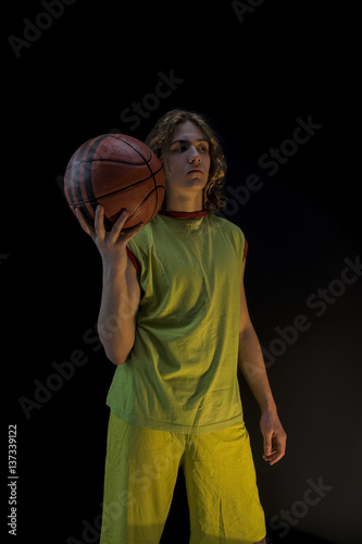 Young boy with long blond hair wearing a green jersey standing and holding up a basket ball