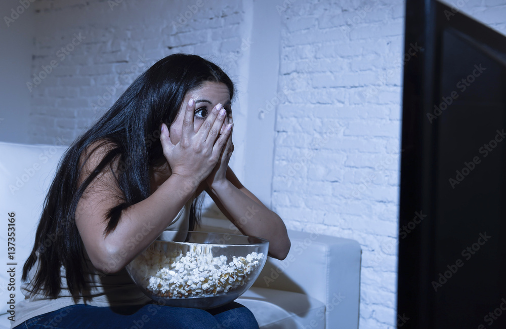 Naklejka premium latin woman at home sofa couch in living room watching television scary horror movie or suspense thriller