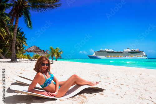 Cruise vacation concept. Cruise ship in the sea near the tropical island with woman lying on a beach bed near the sea. Relaxing and enjoying on vacation.