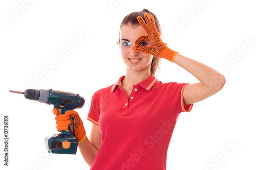 beautiful young slim builder girl in glasses makes renovations with drill in her hands shows OK isolated on white background