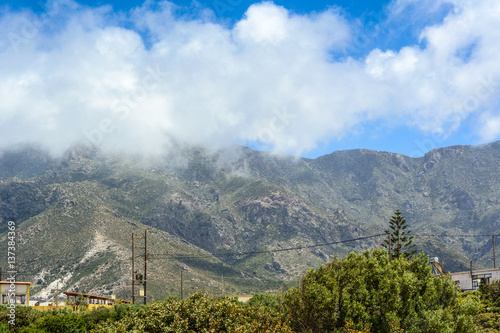 Mountainous nature of Crete Island in Greece. Low clouds hide tops of mountains under blue sky. Houses and typical wooden electric poles with wires under voltage.