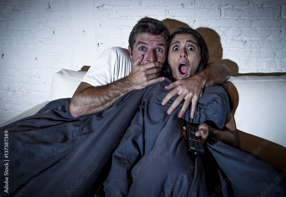 Naklejka premium attractive couple at home enjoying watching television horror movie covering with blanket