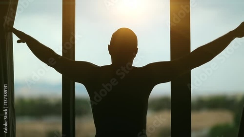 Man opens the curtains in the morning and looks out the window. Young man opens big window curtains and lets the sun light in the room