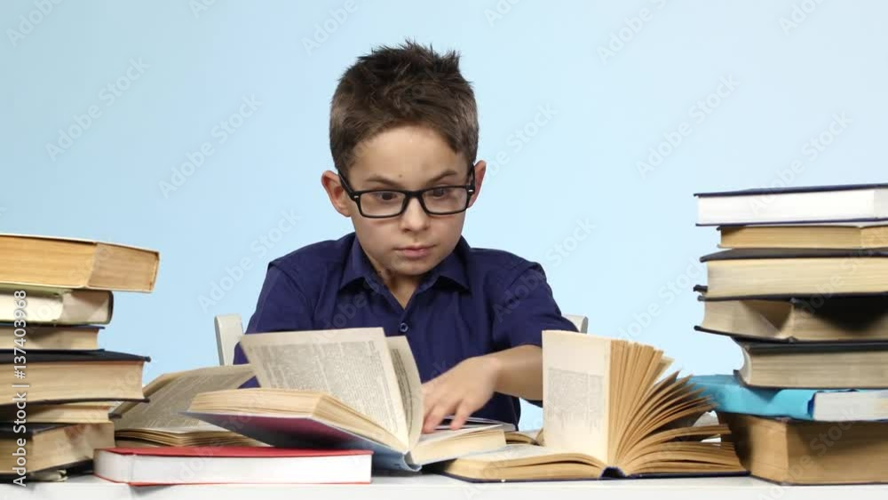Boy with glasses sits at a table and excitedly that is looking for. Blue background. Boy with glasses sits at a table and excitedly that is looking for. Blue background.