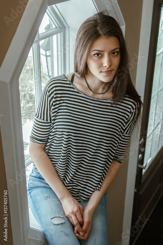 Closeup portrait of pensive sad Caucasian young beautiful woman model with messy long hair in ripped blue jeans and striped t-shirt sitting indoor by window, looking in camera, toned with filters.