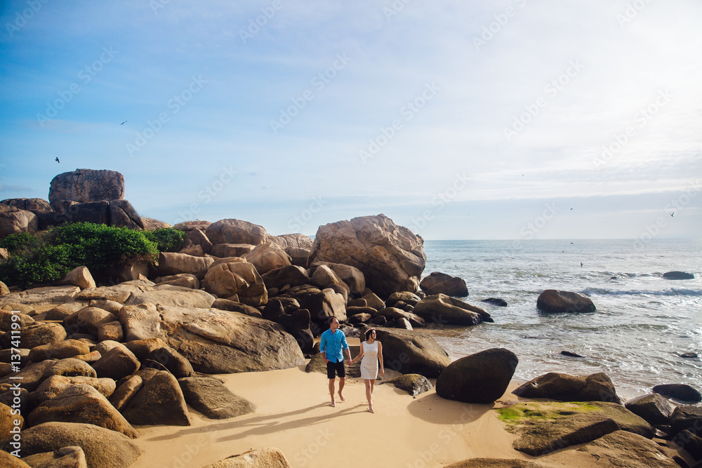 Young couple in love holding hands, walking on a sea beach in summer. The man in the blue shirt and the girl in a white dress with a wreath in her hair Young couple in love holding hands, walking on a sea beach in summer. The man in the blue shirt and the girl in a white dress with a wreath in her hair