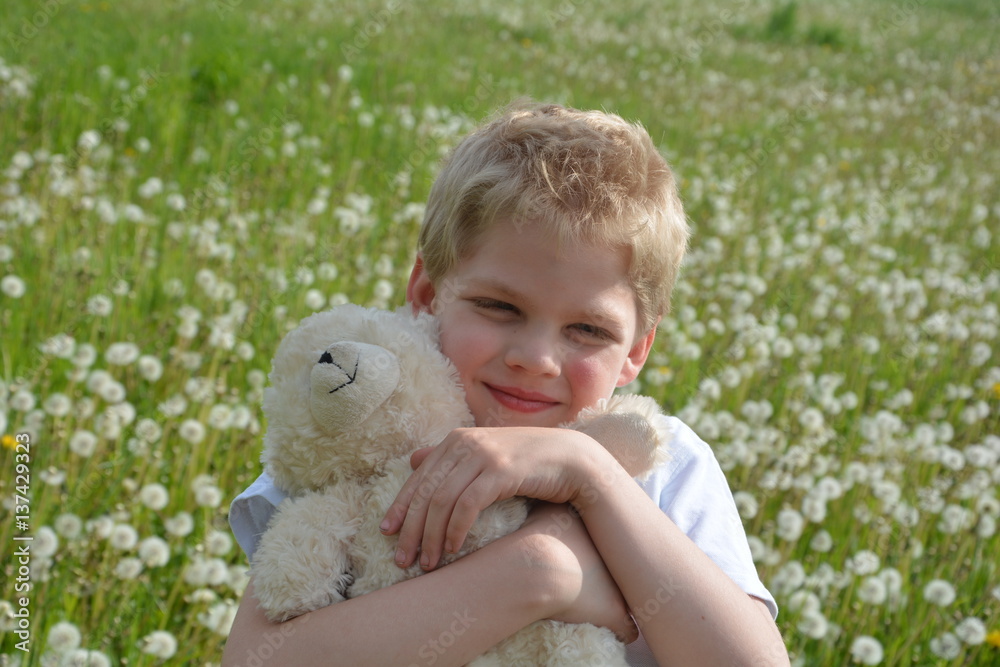 Kleiner blonder Junge auf Wiese mit Pusteblumen, hält weißen Teddybär fest im Arm und schaut nach vorne
Kleiner blonder Junge auf Wiese mit Pusteblumen, hält weißen Teddybär fest im Arm und schaut nach vorne