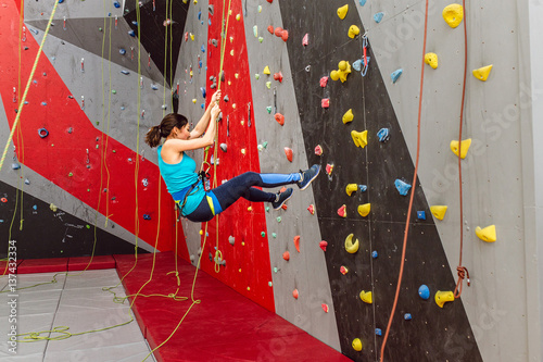 Happy sporty woman with climbing equipment hanging on a rope at indoor rock-climbing artificial wall
