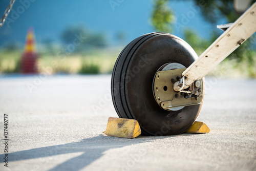 A rear landing gear and wheel chocks of a small aircraft on the ground with blurry nature and traffic cone in the background.