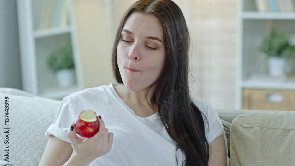 Tilt down of young pregnant woman in pajamas sitting on sofa at home and eating apple Tilt down of young pregnant woman in pajamas sitting on sofa at home and eating apple