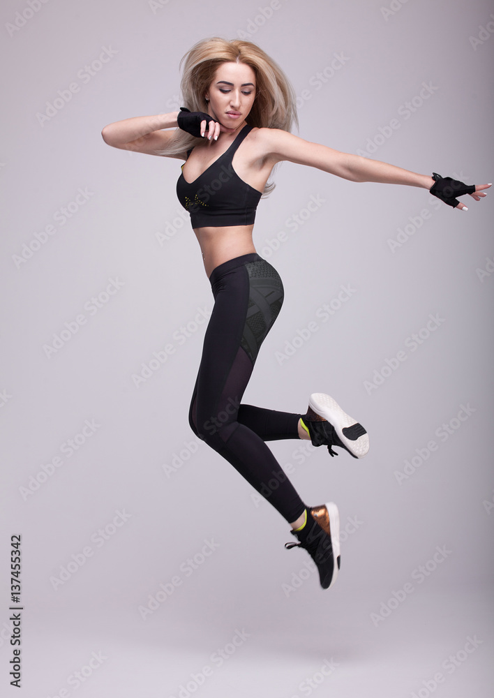 Beautiful young blonde fit woman with long hair, happily jumping on a grey background. Slim woman in sport clothes.