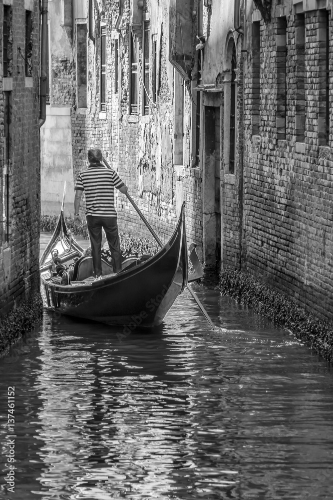 Fototapeta premium Venetian gondolier navigates with his gondola in a narrow canal in the historic center of Venice, Italy. in black and white