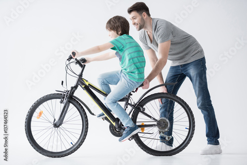 side view of father helping son to ride bicycle on white