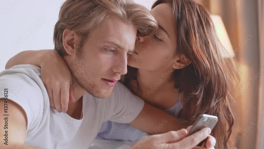 Beautiful Caucasian female kissing her boyfriend while he is concentrated on mobile phone sitting in bed in the morning in slowmotion Beautiful Caucasian female kissing her boyfriend while he is concentrated on mobile phone sitting in bed in the morning in slowmotion