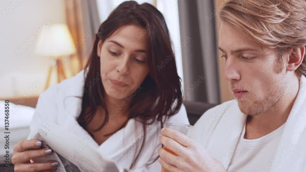 Young Caucasian pair of lovers in white bathrobes reading morning newspaper drinking juice from glass and smiling laughing in slowmotion Young Caucasian pair of lovers in white bathrobes reading morning newspaper drinking juice from glass and smiling laughing in slowmotion