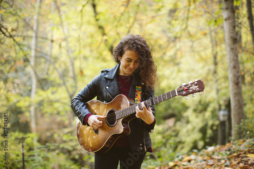 Beautiful young woman playing guitar on forest, fashion lifestyle. Girl wearing black jacket.