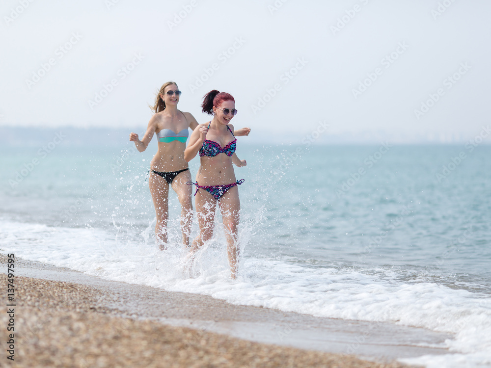 Two girls in bikini running along seashore splashing sea water