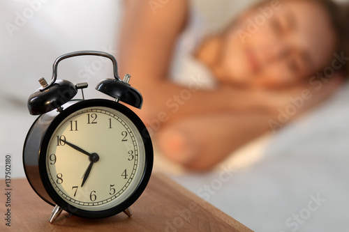Beautiful young woman sleeping and smiling while lying in bed comfortably and blissfully on the background of alarm clock is going to ring. Sunbeam dawn on her face.