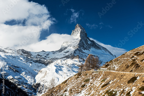 Scenic winter moody view on snowy Matterhorn peak with pine trees, bright blue sky with white clouds, snowy mountains and mountain trail in Switzerland. photo