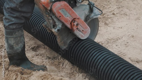 Warker nagging black ribbed plastic pipe with circular saw on sand surface, slowmotion photo