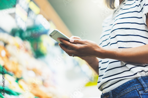 Young woman shopping healthy food in supermarket blur background. Female hands buy products tomato using smartphone in store. Hipster at grocery using smartphone. Person comparing price of produce