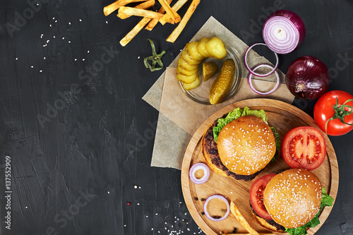 Craft beef burgers on round wooden cutting board with vegetables and french fries. Flat lay on black textured background.