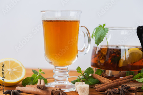 Clear glass and teapot of hot black herbal tea with lemon, mint and Melissa leaves on light rustic wooden table. Summer, Autumn, winter drink. Cinnamon and cane sugar for decoration. Top view.
