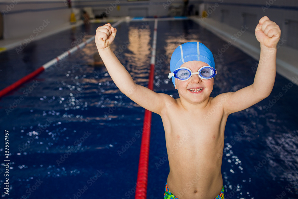 Boy swimmer enjoys, raising both hands up, behind his swimming pool. Gesture of winner