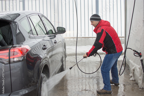 A man in red jacket washing his car in self-service station with high pressure blaster.