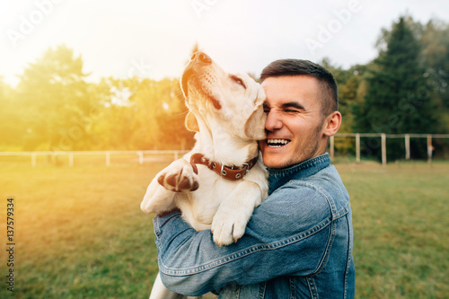 Canvas Print Happy young man holding dog Labrador in hands at sunset outdoors