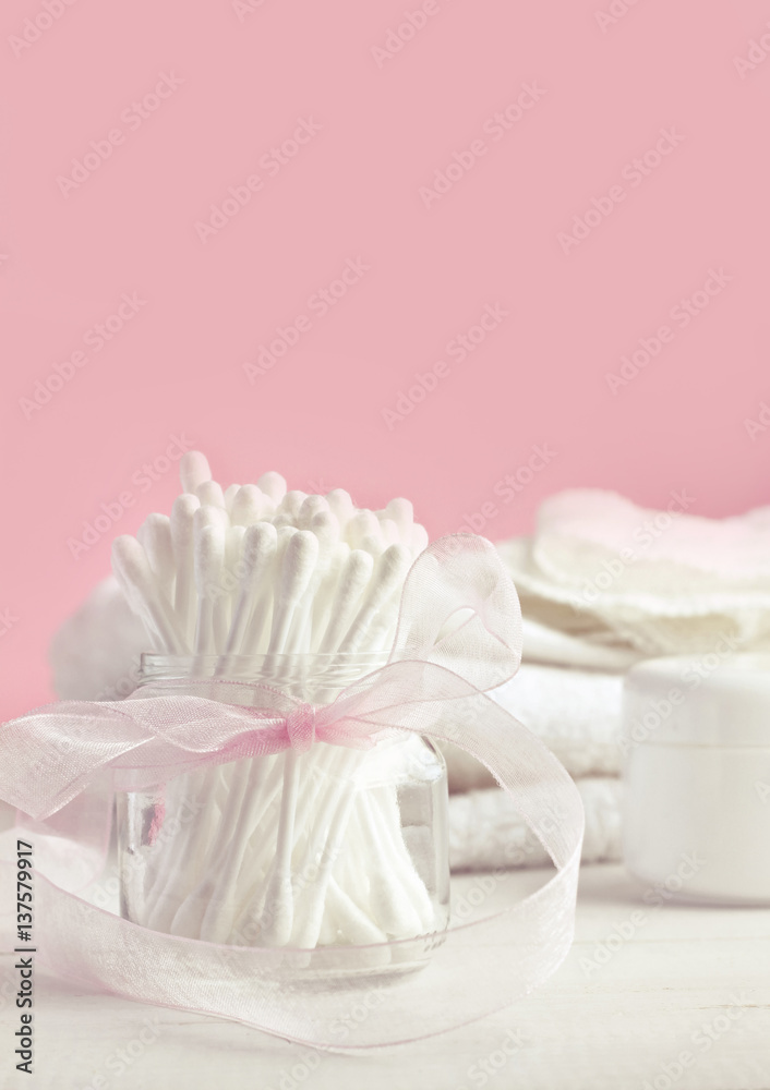 White cotton swabs in glass jar with ribbon bow decor, pink background. Feminine beauty care. Vertical set. Soft light and focus. White cotton swabs in glass jar with ribbon bow decor, pink background. Feminine beauty care. Vertical set. Soft light and focus.