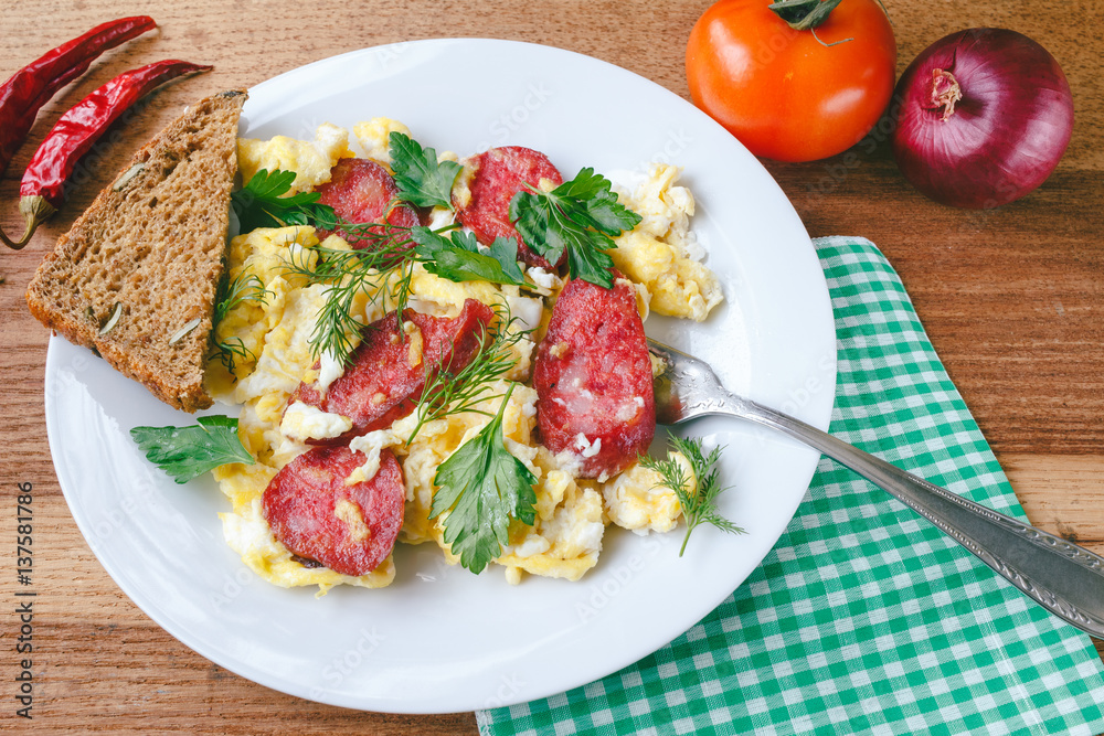 Fresh cooked scrambled eggs with sausage and herbs in white plate.Bread, napkin, fork, vegetables on wooden board. Fresh cooked scrambled eggs with sausage and herbs in white plate.Bread, napkin, fork, vegetables on wooden board.
