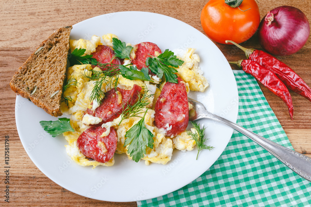 Fresh cooked scrambled eggs with sausage and herbs in white plate.Bread, napkin, fork, vegetables on wooden board. Fresh cooked scrambled eggs with sausage and herbs in white plate.Bread, napkin, fork, vegetables on wooden board.