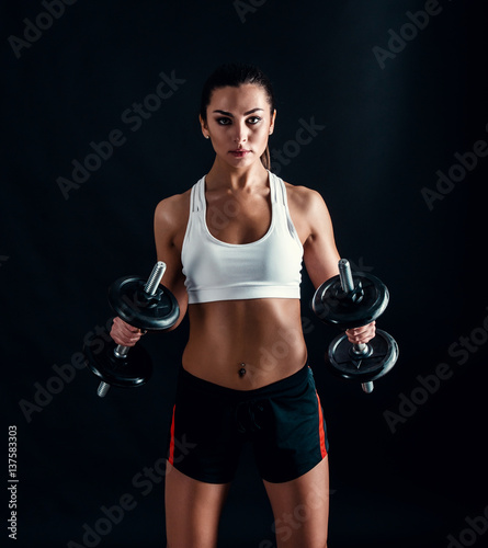 Athletic young woman doing a fitness workout against black background. Attractive fitness girl pumping up muscles with dumbbells.