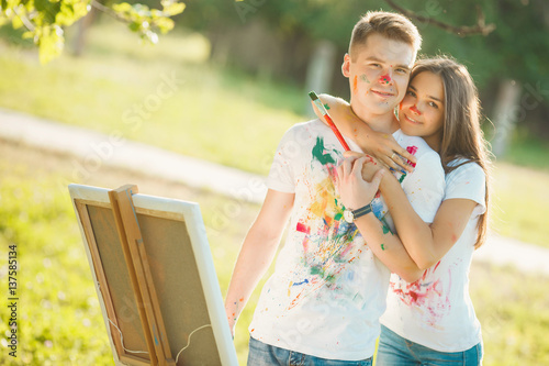 Young beautiful woman embracing her boyfriend. Pretty young guys looking at camera and smiling near the easel outdoors, their t-shirts colorised by themselves. photo