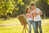 Pretty couple having fun at open air outdoors. Young man and woman drawing by colorful paints on each other`s t-shirts with brushes near the easel on the nature.