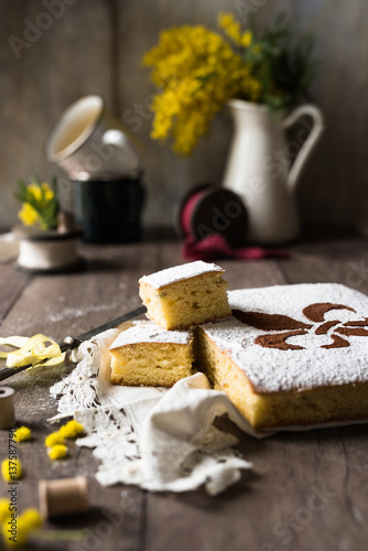 fette di torta gialla allo zafferano posate su pizzo antico e base rustica di legno; fiori di mimosa sfuocati sullo sfondo