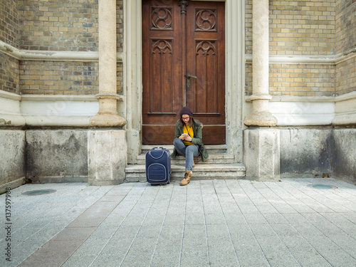 Young hipster woman is sitting on the stairs of the building in old town using her mobile phone and having a suitcase next to her.