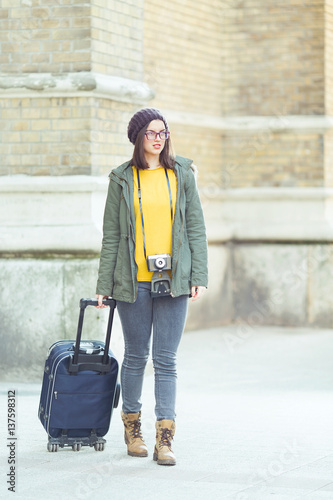 Smiling young hipster woman walking down the street of old town with a suitcase and vintage camera