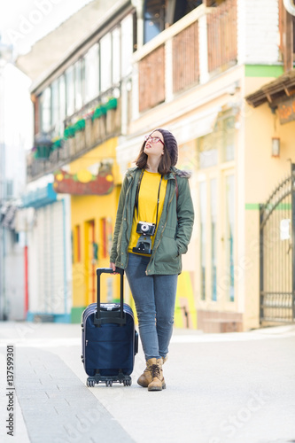 Smiling young hipster woman sightseeing while walking down the street of old town with a suitcase and vintage camera