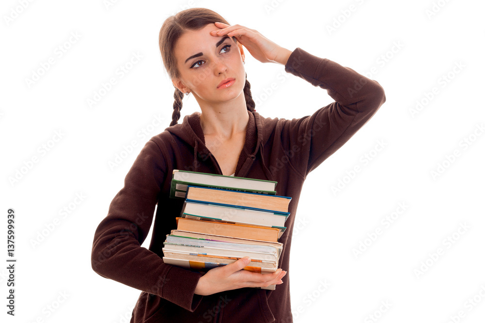 tired student girl in brown sport clothes with a lot of books in her one hand and second on her head posing isolated on white background tired student girl in brown sport clothes with a lot of books in her one hand and second on her head posing isolated on white background