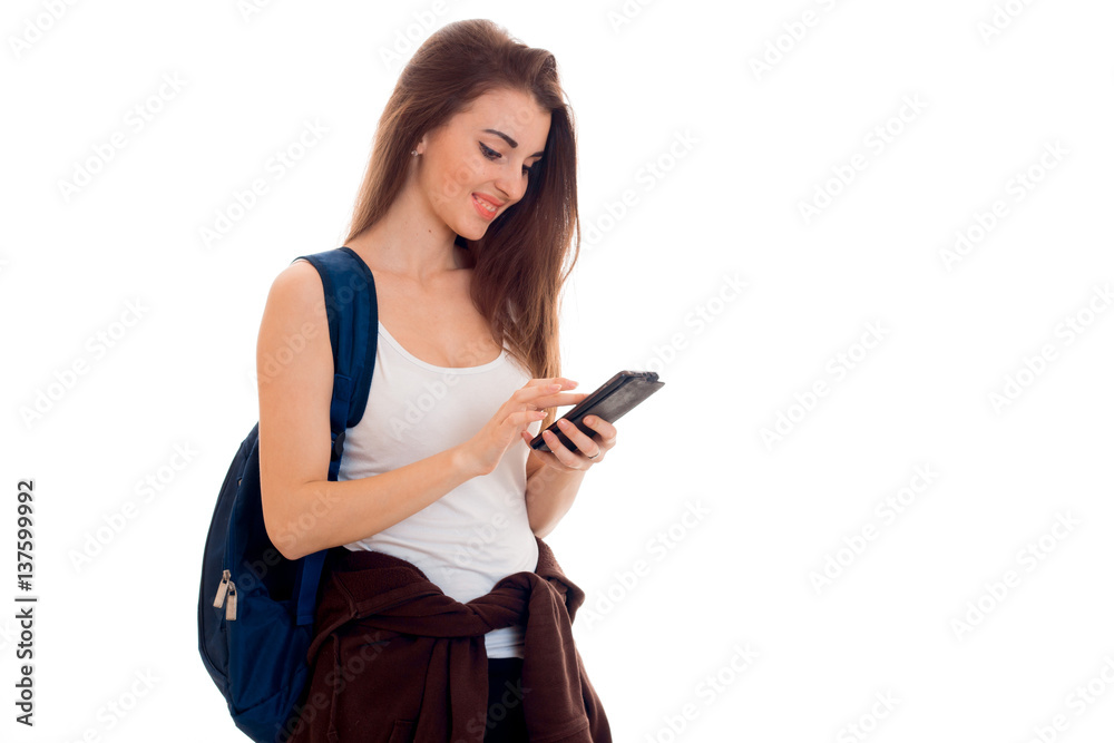 cheerful smart student girl in brown sport clothes with backpack on her shoulders looking at her mobile phone and smiling isolated on white background cheerful smart student girl in brown sport clothes with backpack on her shoulders looking at her mobile phone and smiling isolated on white background