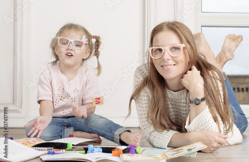 The daughter and mother sit on the floor and draw. The woman helps to the child to draw with markers. 