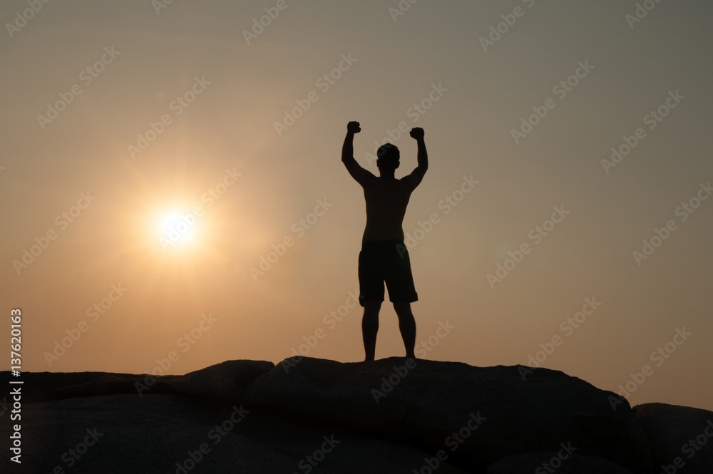 Positive male holding hand up and expressing gladness while standing on stone in Sunset.