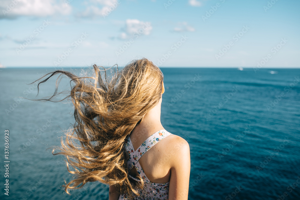 young pretty girl looks at the sea in the summer with her back. Strong winds shook hair