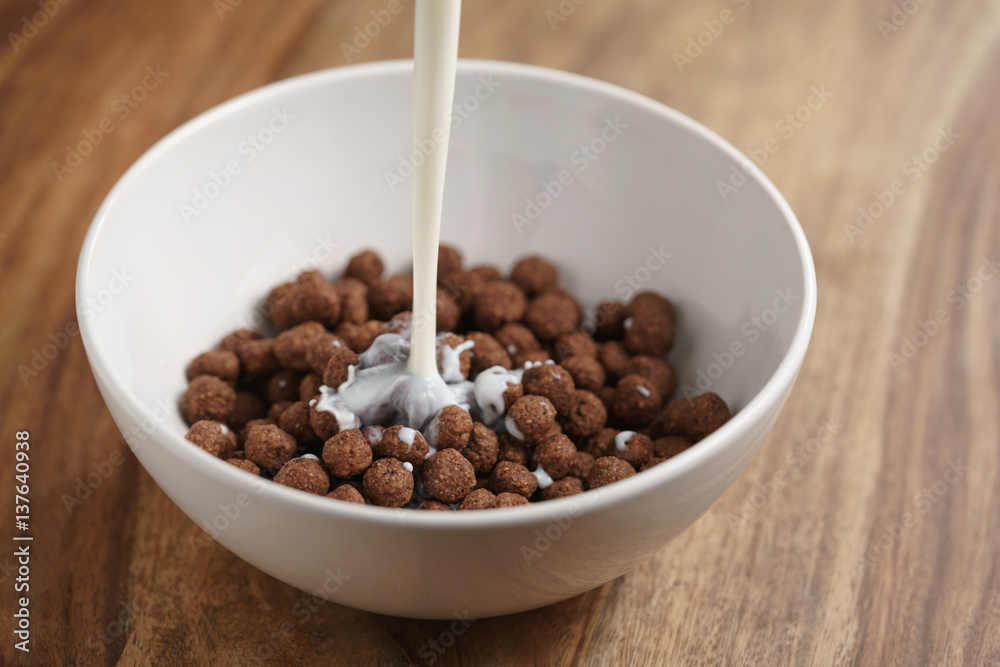 pouring milk into chocolate cereal balls in white bowl for breakfast on wooden table pouring milk into chocolate cereal balls in white bowl for breakfast on wooden table