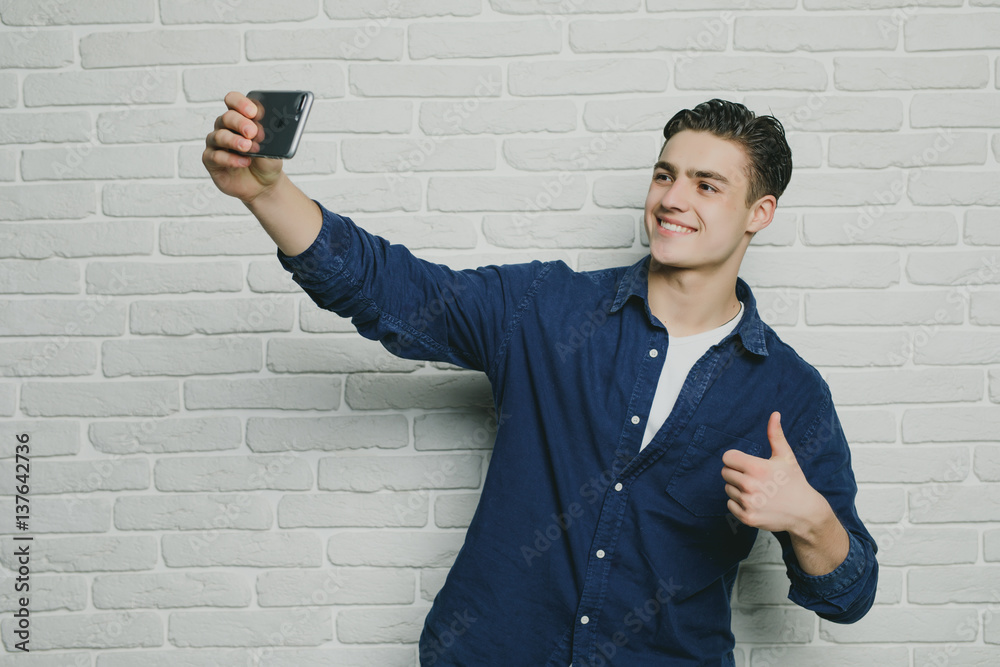 Beautiful young man in casual clothes make selfie on a background of white brick wall Beautiful young man in casual clothes make selfie on a background of white brick wall