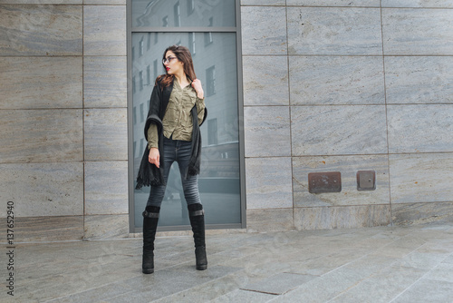 Fashion consept: beatiful young girl with long hair, glasses, red lips standing near modern wall wearing in green suit and grey jeans. She wanted talking on phone.