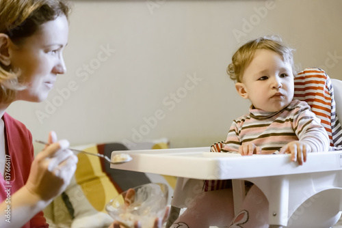 Baby sitting on the highchair and his mother is feeding from the spoon photo